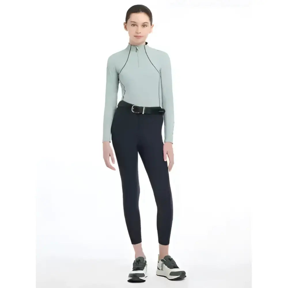 The lemieux young rider model wears a light sage green athletic top with black zippers paired with winter breggings navy