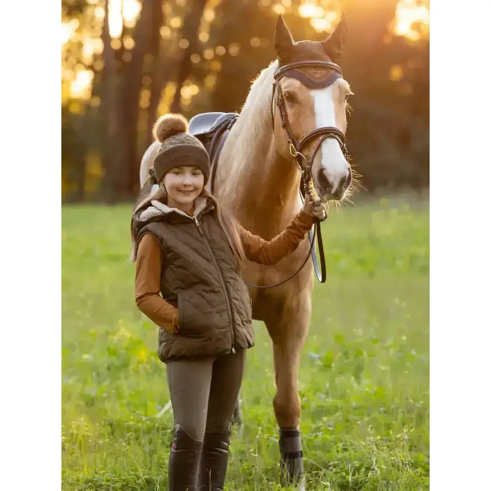 A light brown horse with a white blaze wearing lemieux young rider eloise reversible winter gilet alpine in a field