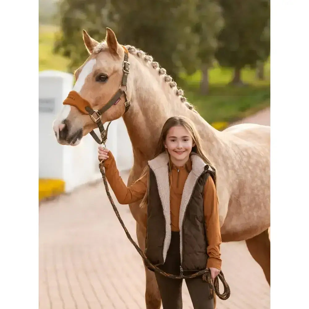 A young girl wearing a brown and white shearling-lined lemieux young rider eloise reversible winter gilet alpine stands by a tan horse
