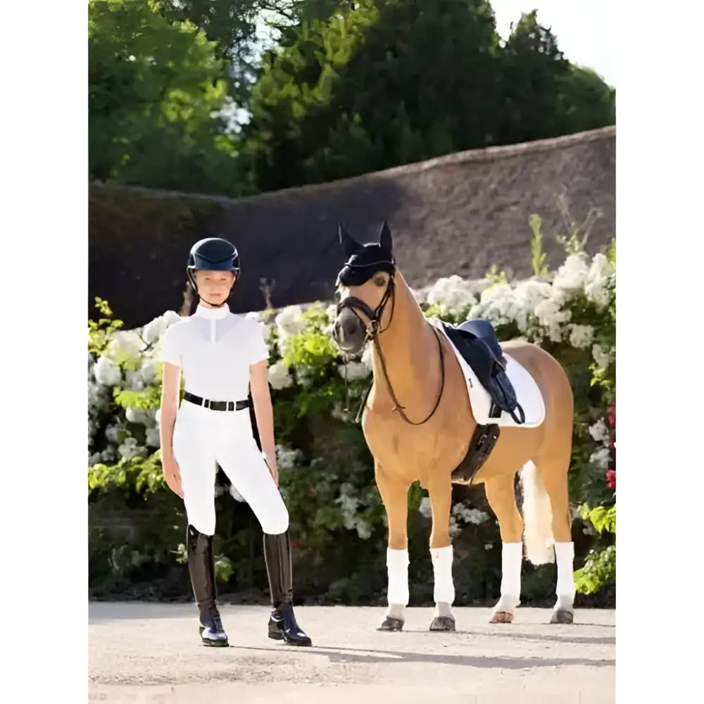 A young woman wearing lemieux young rider eva short sleeve white show shirt stands next to a horse in tack