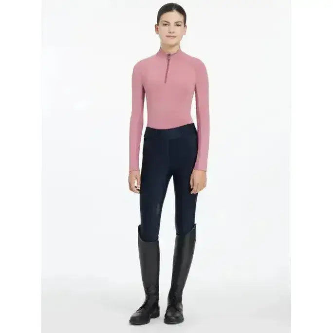 A woman wears the lemieux young rider hope lightweight base layer peony pink equestrian top with black riding pants and boots