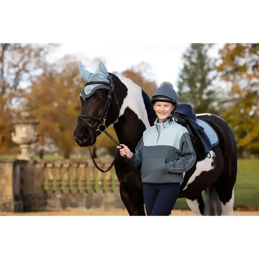 A young girl wearing a lemieux young rider kate quarter zip sweat petrol light blue jacket stands beside a black horse