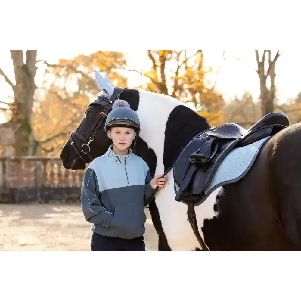 A young rider wearing a lemieux young rider kate quarter zip zip sweat petrol jacket stands beside a pinto horse