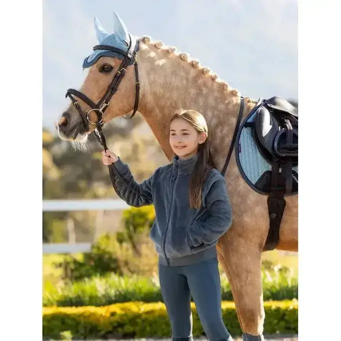 A young girl wearing lemieux young rider libby fleece petrol navy blue fleece stands next to a horse with matching blue tack