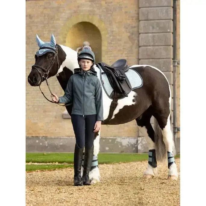 A young rider wearing a grey lemieux young rider lottie soft shell jacket petrol stands beside a brown and white horse