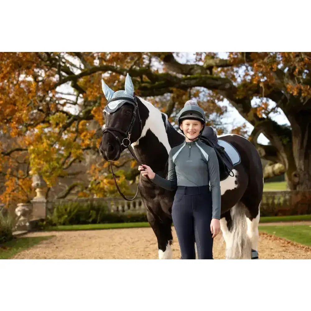 A black and white pinto horse wearing a silver mesh ear cover with lemieux young rider mia mesh base layer petrol