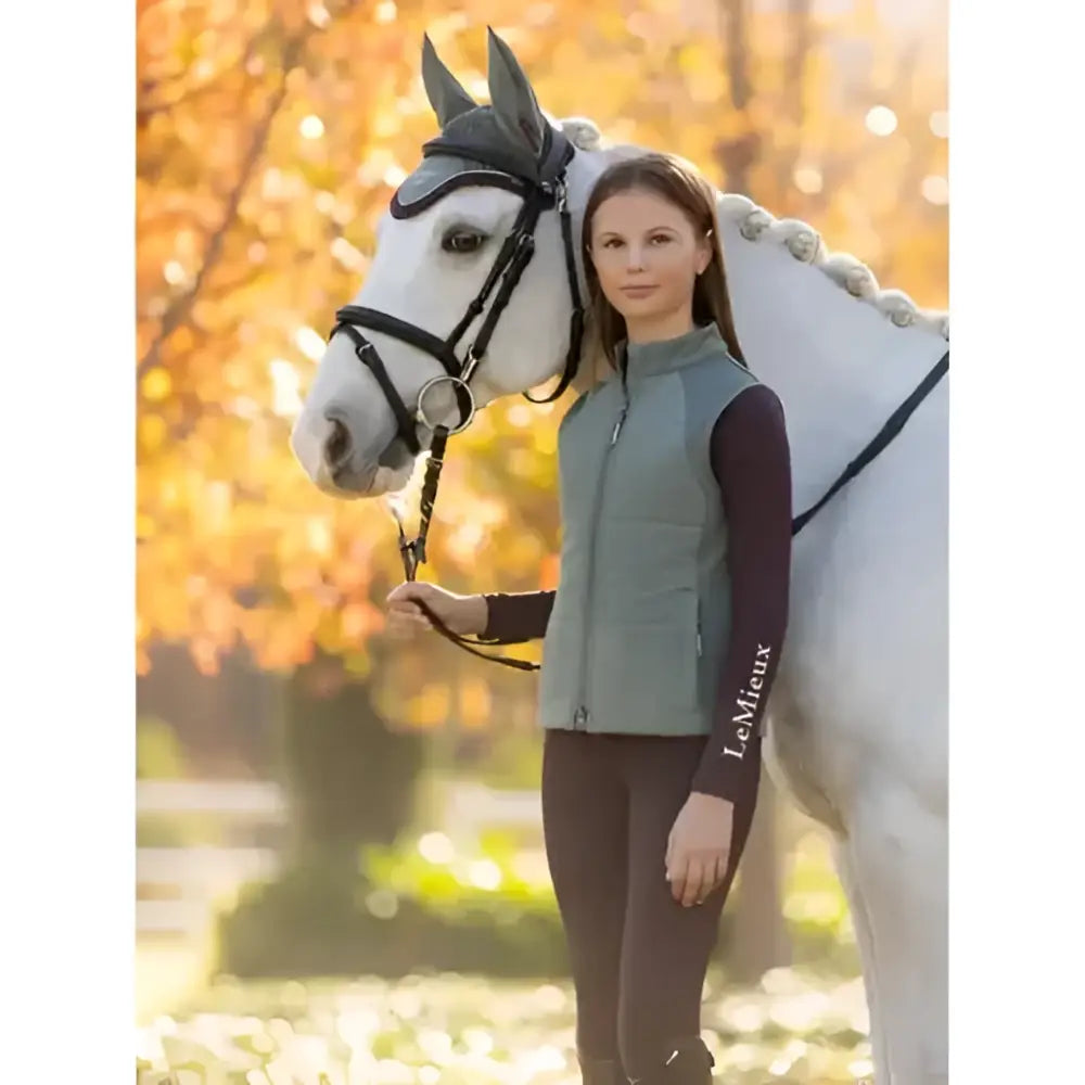 Woman wearing a sage green lemieux young rider quilted vest with black sleeves and the lemieux logo on the left sleeve
