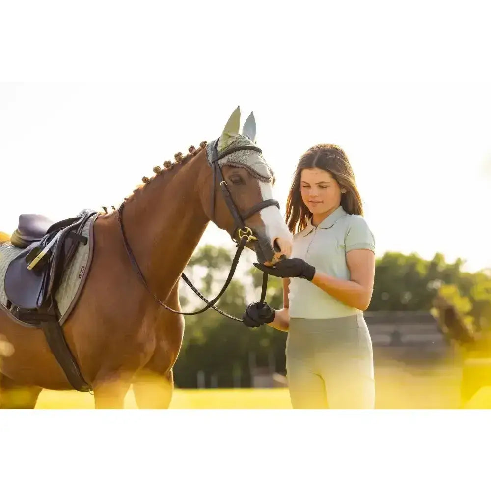 Brown horse with white blaze wearing black bridle next to lemieux young rider polo shirt pistachio in gray saddle pad