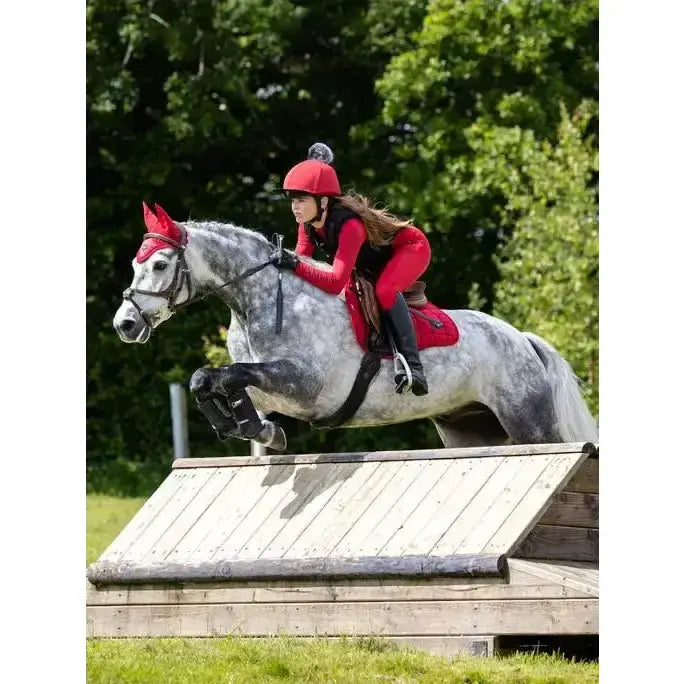 A rider in a red helmet wearing lemieux young rider breech chilli guides a dapple grey horse over a wooden jump
