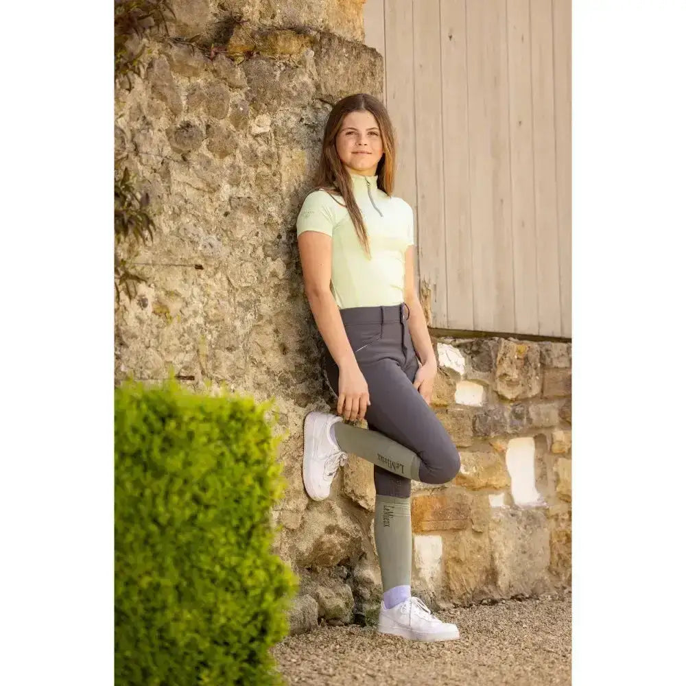 A girl wearing a lemieux young rider short sleeve base layer pistachio in light green with dark gray pants and white sneakers