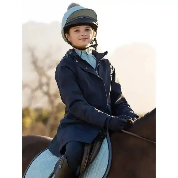 A young girl wearing a navy lemieux young rider skye waterproof coat and matching gloves seated on a horse