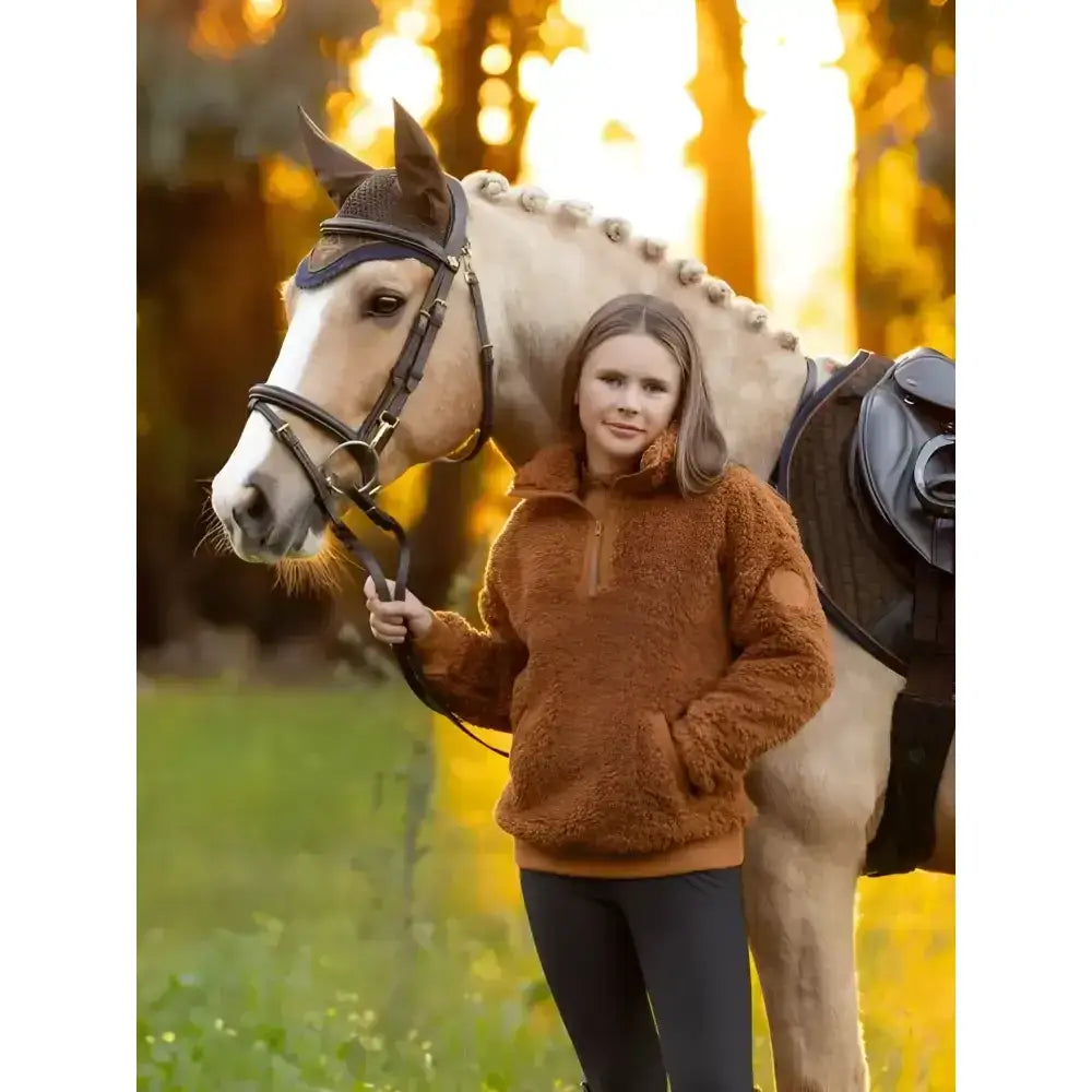 A young woman wears a rust-colored lemieux young rider tia teddy fleece pullover standing next to a light cream horse