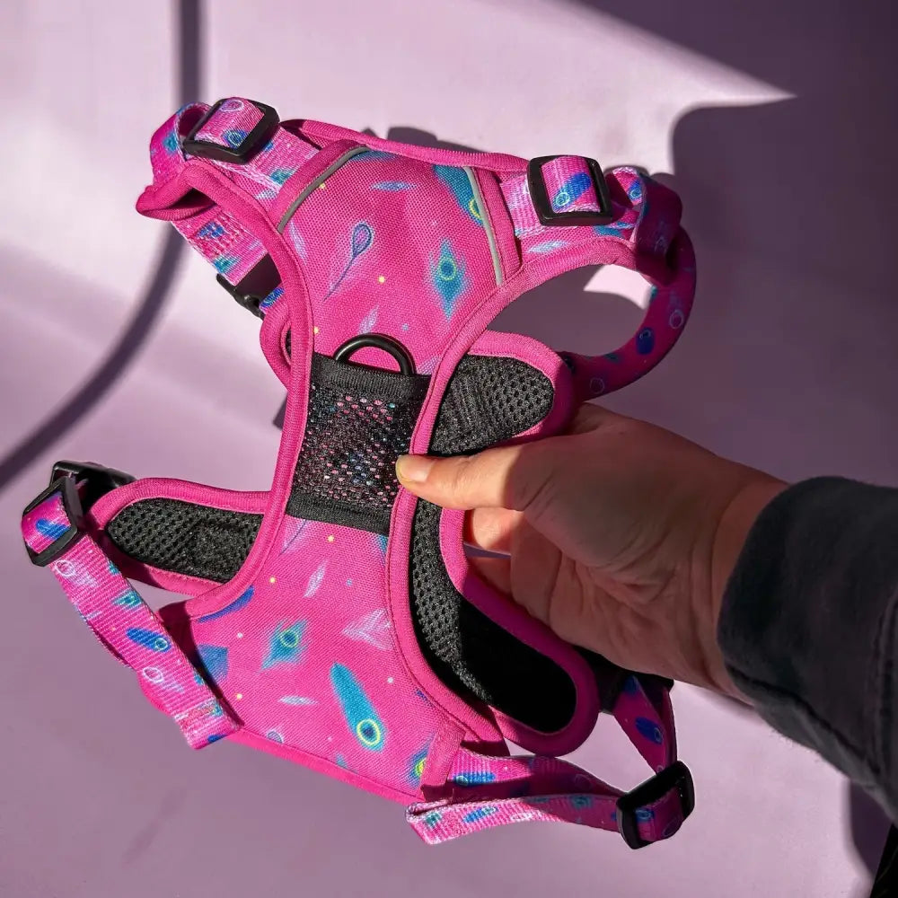A vibrant pink tactical dog harness with peacock feather print, black mesh panels, and adjustable straps for pup chic style