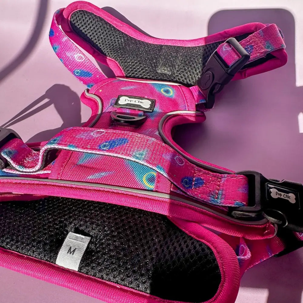 A vibrant pink tactical dog harness with playful peacock feather print, black mesh padding, and adjustable buckles for pup chic