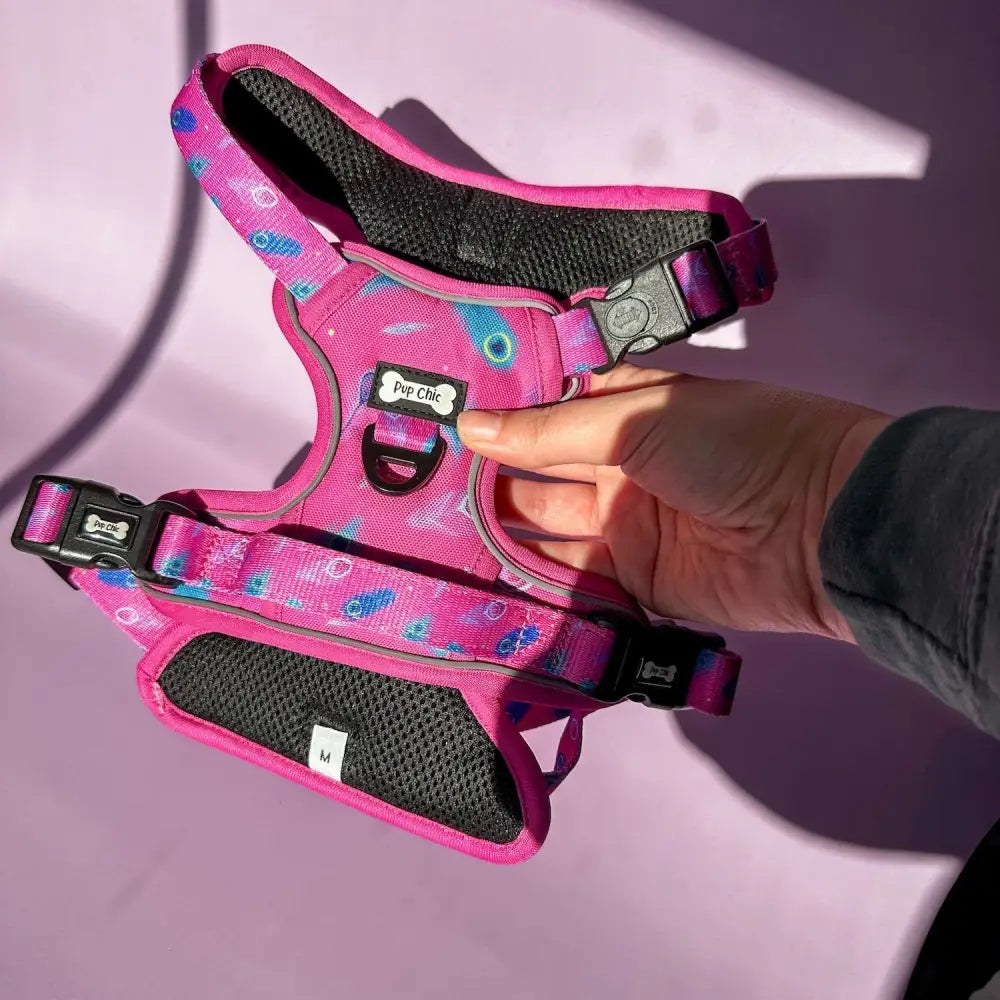 A vibrant pink tactical dog harness with peacock feather print, black mesh padding, adjustable buckles, and pup chic label