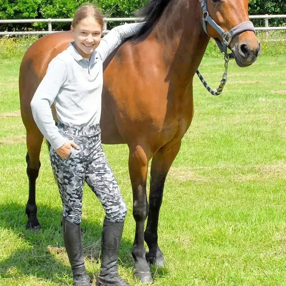 A sleek chestnut horse stands calmly in grass next to rhinegold children’s silicone full seat riding tights in grey camo
