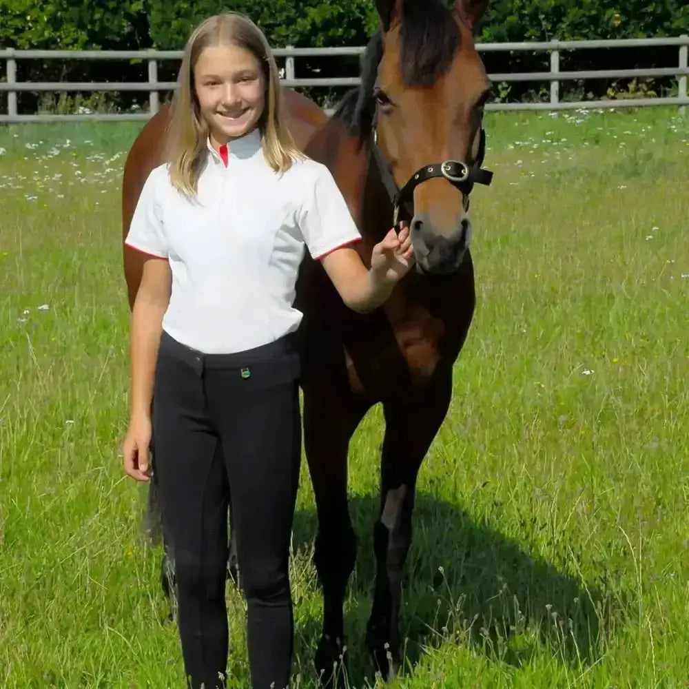 A young woman stands beside a brown horse wearing white and red shirt with black rhinegold childs jodhpurs essential everyday