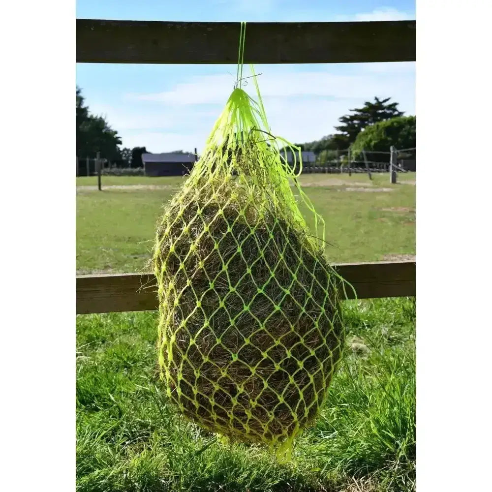 A bright neon yellow mesh rhinegold monarch slow feeder haynet small hole filled with hay hanging on a wooden fence post