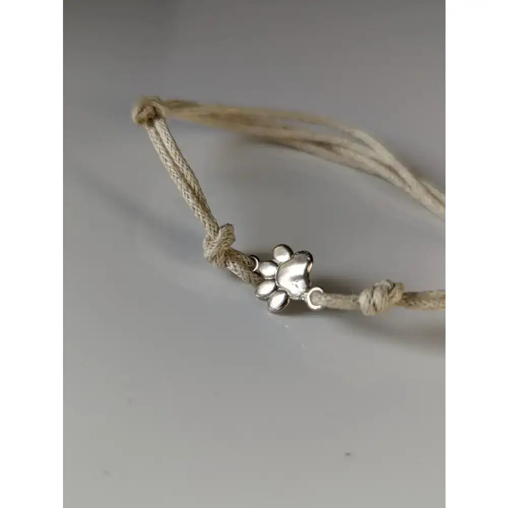 A silver steed paw print charm on a light beige braided cord bracelet featuring a delicate paw print design