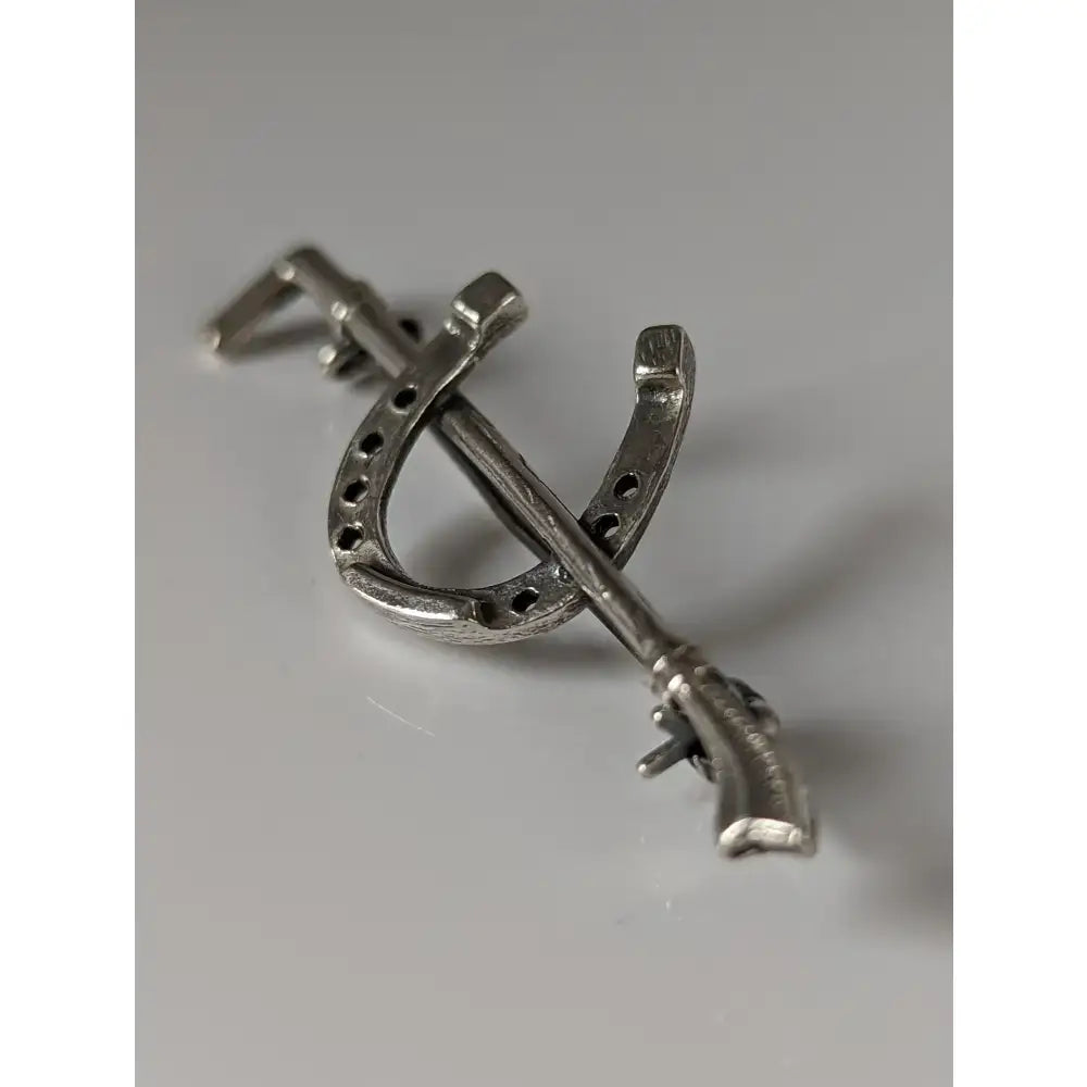 Silver steed shoe stock tie pin featuring a silver metal brooch shaped like a horseshoe crossed with a miniature rifle