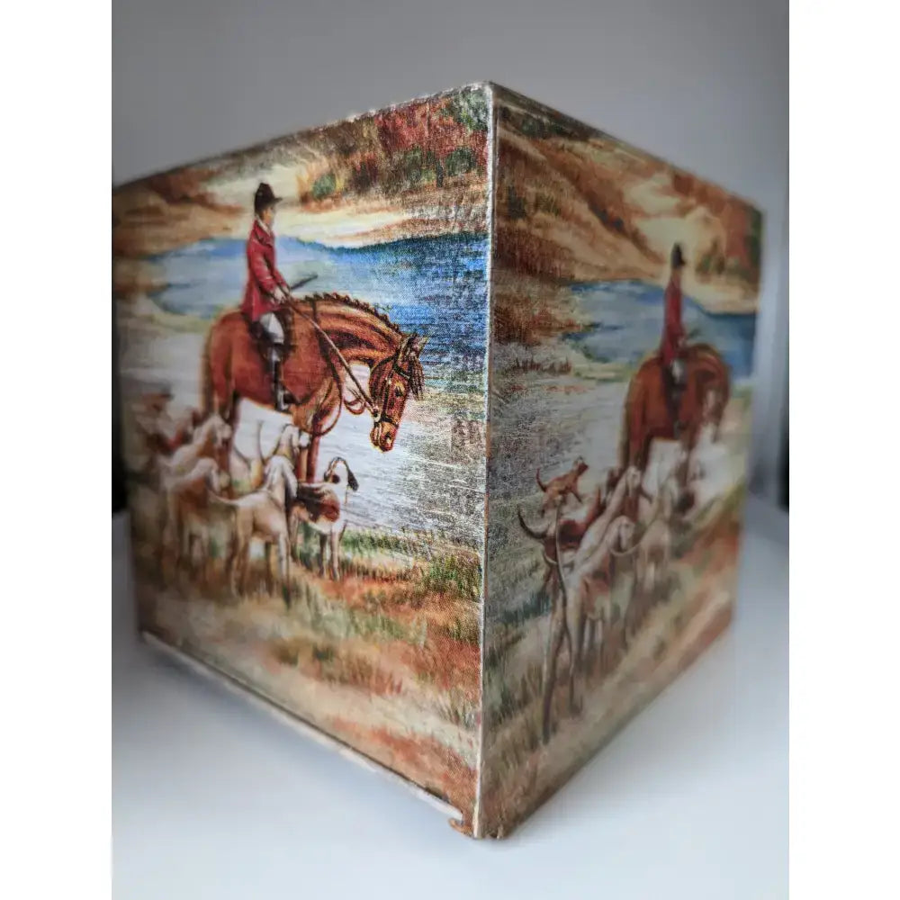 A cube-shaped silver steed tissue box with a painted red riding man on horseback and hounds on its sides
