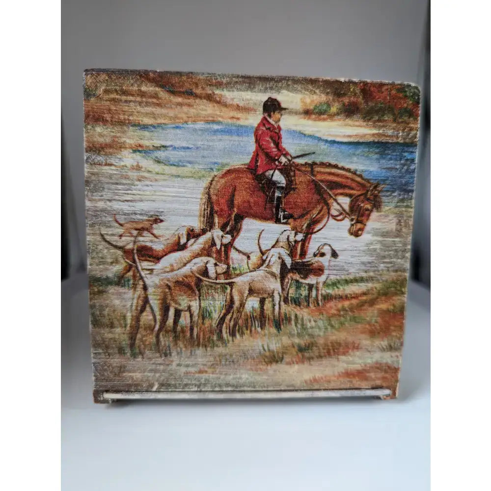 Square wooden silver steed tissue box with painted rider in red atop brown horse and hounds near water scene