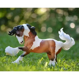 A breyer gypsy vanner model horse with a brown and white pinto coat standing in grass inspired by local traveler communities