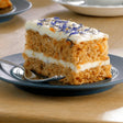 A slice of carrot traycake handmade cake with orange crumb white frosting and purple edible flowers by handmade cake co