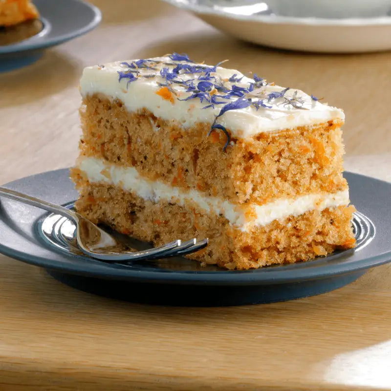 A slice of carrot traycake handmade cake with orange crumb white frosting and purple edible flowers by handmade cake co