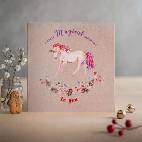 A soft pink christmas card merry featuring a white unicorn with red mane and festive pinecones and candy canes