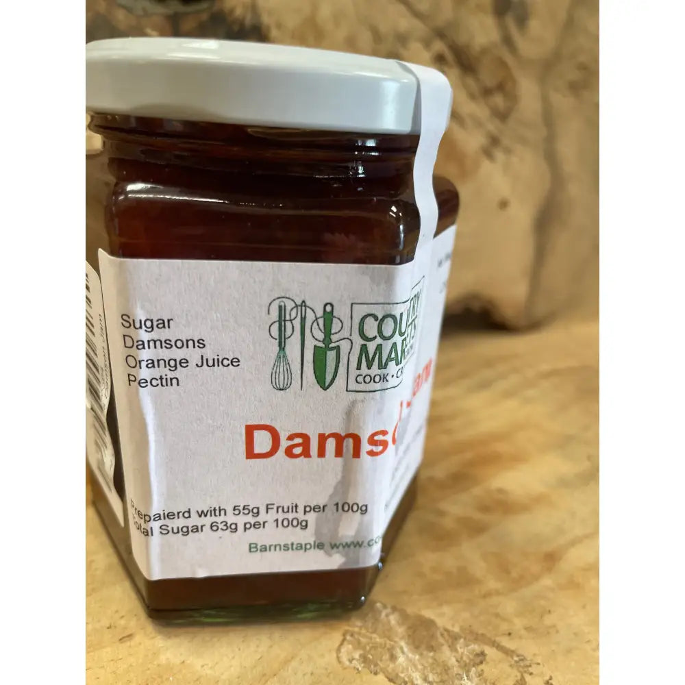 A glass jar of country markets damson jam with a white lid filled with dark reddish-purple preserve
