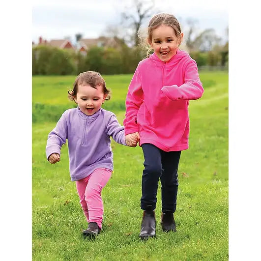 Two girls wearing brightly colored equetech dinky tots jodhpurs running hand in hand through a grassy field