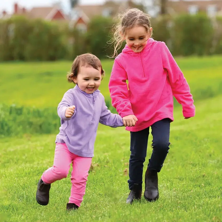 Two kids run hand-in-hand in bright fleece hoodies showing equetech dinky tots jodhpurs in green field