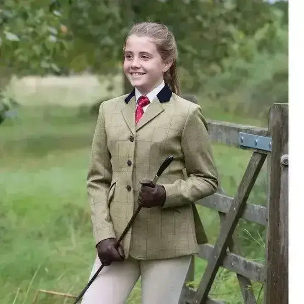 A young girl wears an equetech junior foxbury deluxe tweed riding jacket in beige with black collar trim and brown gloves