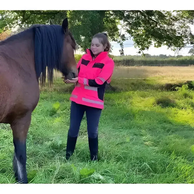 A woman wearing an equisafety inverno childs reflective hi viz riding jacket in bright pink stands in a grassy field