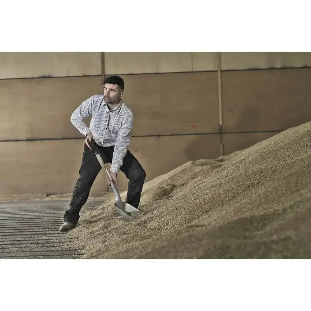 A man wearing a fort tattersall shirt green brushed flannel shirt holds a shovel beside a large grain mound