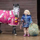 A gray pony wearing a pink blanket with rabbits and carrots stands by a child showing the gallop small haynet for horse feed