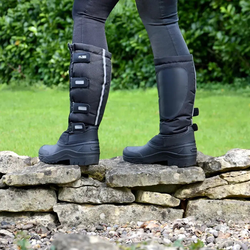 Tall black Equestrian Atlantic Winter Boots with adjustable straps on a stone wall