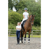 A chestnut horse with black and white leg wraps stands next to a hy equestrian children’s roka regal show shirt in an arena