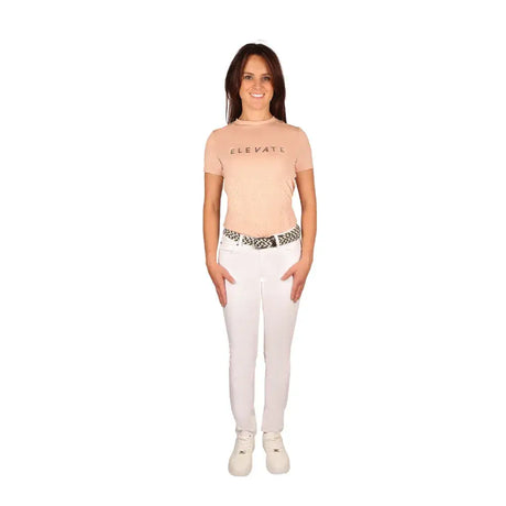 A woman wearing a beige equestrian elevate allure t-shirt with dark lettering paired with white pants and patterned belt