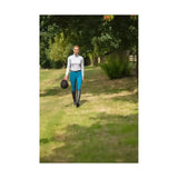 A woman walks holding a black riding helmet wearing hy equestrian hyedition full seat riding breeches on a grassy path
