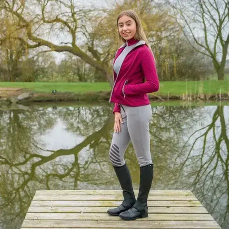 A woman wearing gray fitted equestrian mizs arabella breeches paired with black knee-high boots and a magenta jacket