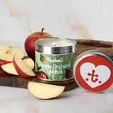 A lit green-labeled apple orchard antics thelwell collection candle in a silver tin with a red heart lid by hy equestrian