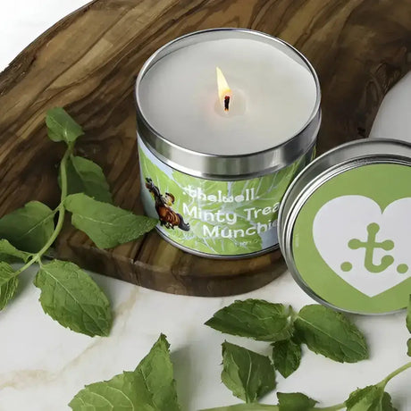 A lit white scented candle in a silver tin with a minty tree munchie label and fresh mint leaves from hy equestrian thelwell collection long-lasting aromatic candle