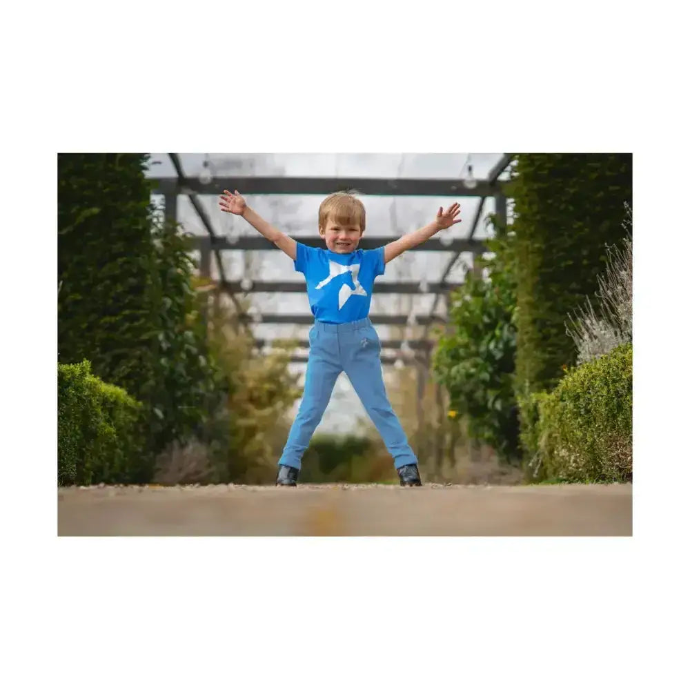 A cheerful boy wearing bright blue pull-on tots jodhpurs from hy equestrian zeddy tots stands outdoors joyfully