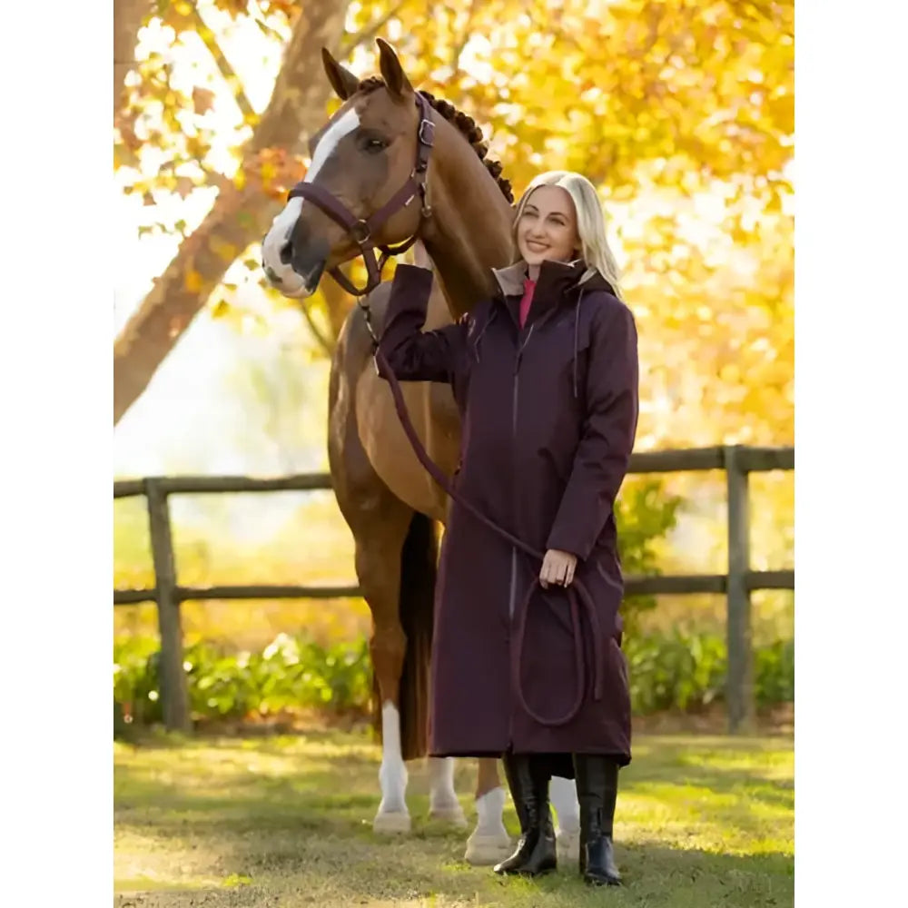 A woman stands beside a brown horse wearing the lemieux all elements robe damson in dark purple hooded riding coat