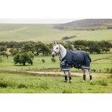 A dapple grey horse wearing a navy lemieux arika storm-tek rug 50g with white trim and a small logo in a green field