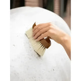 A hand holding the lemieux artisan deep clean dandy brush brown with natural bristles brushing white foam off a horse