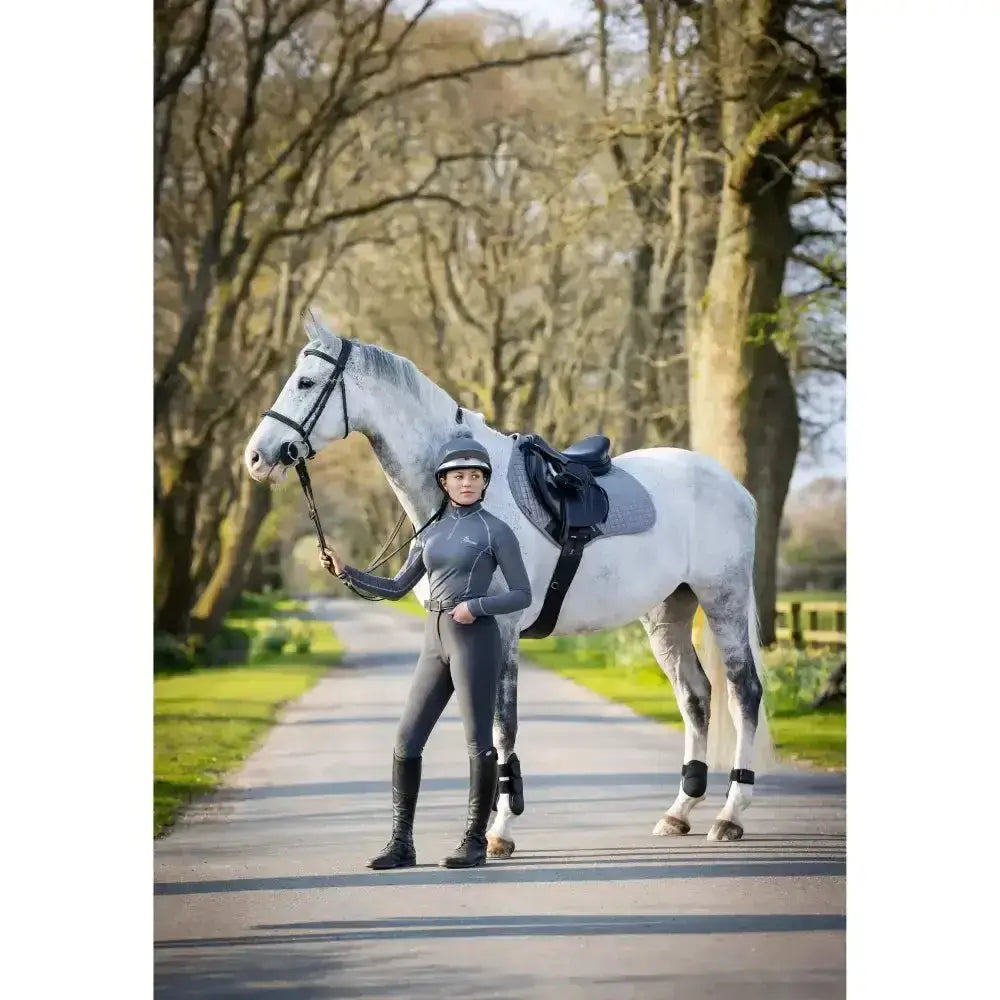 A sleek white horse with black saddle stands beside a rider wearing lemieux base layer slate grey matching heaven gear
