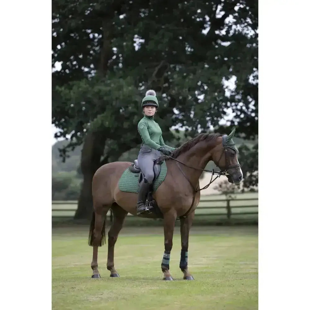 A brown horse wearing lemieux base layers hunter green saddle pad and protective leg wraps in an outdoor field